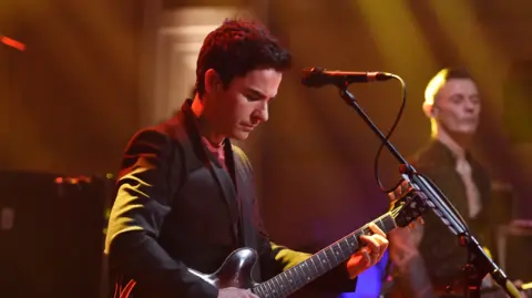 Sarah Jeynes/BBC Lead singer Kelly Jones is playing guitar in front of a microphone. He has short black hair and is wearing a suit jacket.