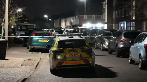 Eddie Mitchell A marked police car is parked on a residential street at night with multiple vehicles on both sides and police tape cordoning off the area.