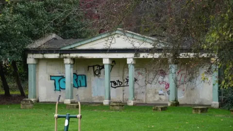 Shaun Whitmore/BBC A pale stone pavilion fronted by six mint green columns. The pavilion is boarded-up, with the boards covered in graffiti. The area in front of the pavilion is laid to lawn.  