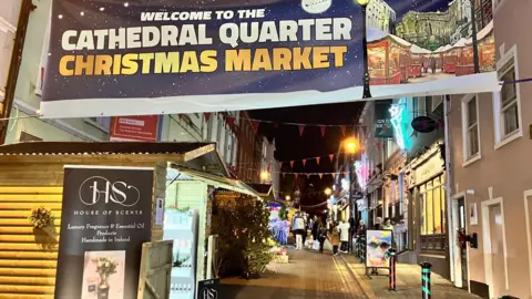 A narrow street with a number of log cabins trading. 
A banner can also be seen which reads: 'Welcome to the Cathedral Quarter Christmas Market'