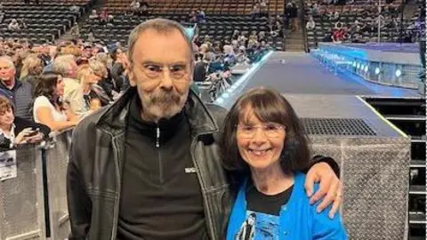 Phil Beard Phil and Eileen pose with their armas around each other near a stage with other fans waiting in the background seats before a performance
