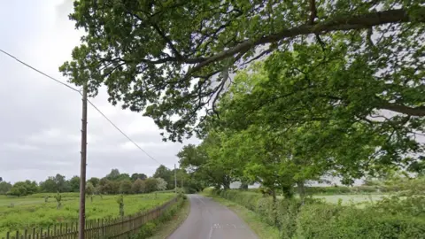 Google A stretch of Packington Lane, near Meriden, a rural area of Warwickshire. A road is surrounded by fields and trees and there is a fence to one side.