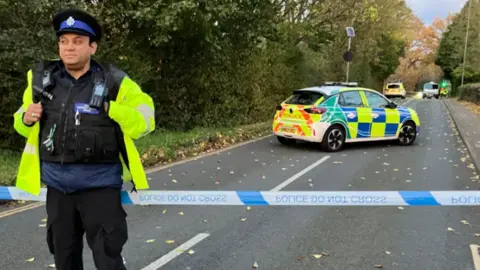 A police officer wearing hi-vis at the cordon to the crash in Lawrence Weston on Sunday. The male police officer is standing in front of police tape. Two police cars are seen within the cordon.