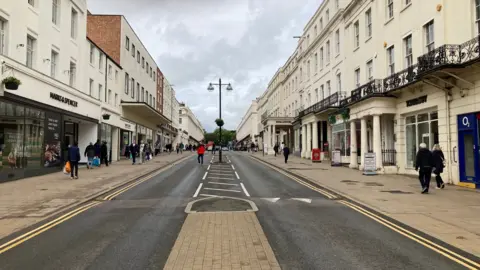 Leamington Spa High street with shops on either side of a long straight road. People can be seen walking on either side with one man in a red coat crossing the road.