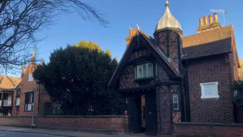 Jon Wright/BBC A Tudor building, with ornate brick and wooden panels, and a turret above one entrance. There is a low brick wall running along the public pavement.