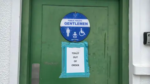 A close up of a green door on a white building. It has a round blue sign on it saying 'Gentlemen' and has in white letters, with white images of a person, a baby and a wheelchair. Underneath it has a a white piece of paper, with 'OUT OF ORDER' in black font, which is taped to the door.