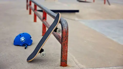 A skate board rested against a metal rail in a skate park. A blue helmet sits on the floor next to it.