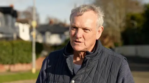 A man standing in the street talking. He is looking just passed the camera. The street is blurred in the background but the sun is illuminating the street, where the outline of grass and white houses can be made out. The man has short grey hair and is wearing a quilted black body warmer.