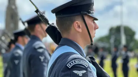 A close-up of a member of the King's Colour Squadron in a line. He has RAF regiment on a badge on his shoulder, he is wearing an RAF cap and had a rifle over his shoulder,