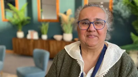 Suffolk High Sheriff Gulshan Kayembe stands looking at the camera, there is a cafe background