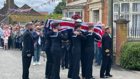 Pallbearers in black Royal Navy uniforms carry the coffin of Matthew Flinders, which is draped in a Union flag with a floral wreath on top. In the background, a crowd watches beneath red and blue bunting.on top with three Royal Navy men on each side and a crowd with punting in the background