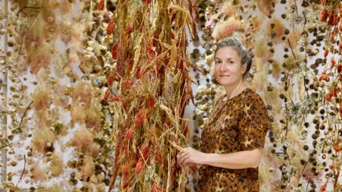 Stockton Council Artist Rebecca Louise Law standing beside red and yellow flowers suspended from the ceiling, Her grey hair is pulled back and she is wearing an orange top. She has blue eyes and is smiling slightly at the camera. 