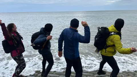 Eva Kriki Runners Chris Parr, Paul Baxter, Eva Kriki and Behzaad Ghouse stand together at the shoreline. They are wearing trial running equipment and clothing of various bright colours and are throwing a pebble into the sea.