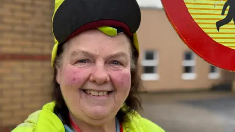 A headshot of Carol Hawke, smiling in her yellow lollipop lady uniform and holding the stop sign.