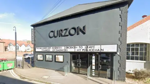 the outside of Canterbury's Curzon cinema. The building is one storey and is dark grey with the word CURZON written on the top half of the wall. There is a white sign horizontal on the front of the building with black lettering of the film listings