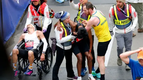 Reuters Four members of medical staff assist exhausted runners at a marathon finish line; one runner in a white vest (Robson) sits slumped in a wheelchair while others support Ajay who is leaning forward, Aaron is stood upright holding him up.