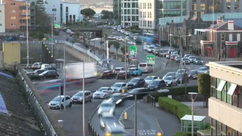 View of busy roads taken from a timelapse film so many of the cars on the roads are slightly blurred showing movement. The lighting is warm suggesting the image has been taken late in the day and vehicle headlights and rear lights are on. This is a wide view showing a number of different roads with traffic going in different directions. High rise and low buildings are visible in the foreground and background.