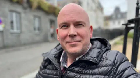Marcus Saul is smiling at the camera as he sits outdoors. Blurred in the background behind him are stone buildings and a road. There are also thin black iron posts just behind him which may be part of the bench he is sitting on. Mr Saul is wearing a black puffer jackets and he has a shaven head, slight beard and blue eyes.