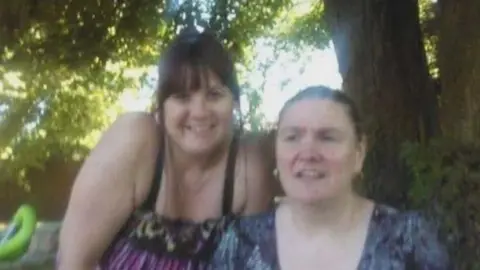 Karen Bassett One woman leans in over a second woman who is sat beside a tree. The leaning woman has tied back dark hair and is wearing a top with black straps and a pink pattern on. The second woman who is sitting is looking off camera and is wearing a floral blue top with tied back dark hair.