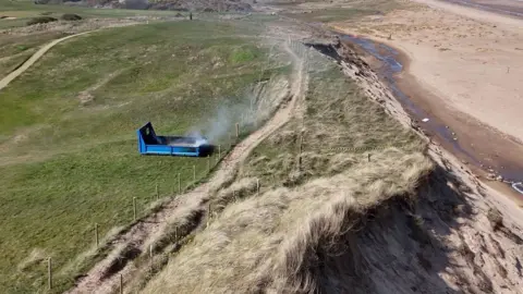 Smoke coming from small blue cordoned off structure on a grassy beach cliff. On the right hand side of the photo you can see sand.