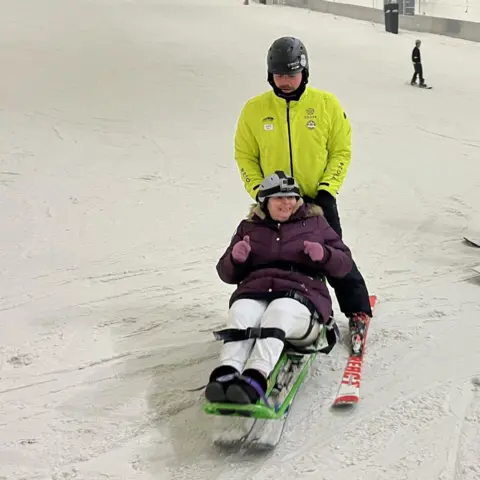 A woman on a sit-ski - an adapted ski with a seat - is guided down a snow slope by a male guide on skis. The woman wears a purple winter coat, purple gloves, white trousers and a grey helmet. The guide wears a hi-vis yellow jacket and black helmet. A snowboarder can be seen in the background.