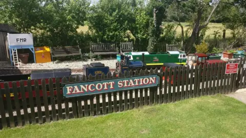 Fancott Miniature Railway Fancott Miniature Railway, with trains and seats on a small track behind a picket fence, which has a "Fancott Station" sign on it.
