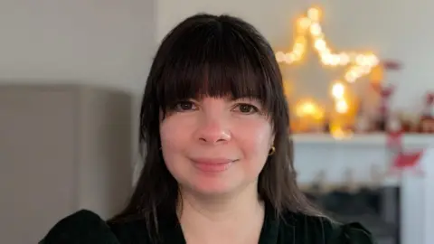 TRISH MCDONOUGH Trish, a woman with brown hair, has a fringe and wears a black long-sleeve top. She smiles in front of an illuminated star in the background.