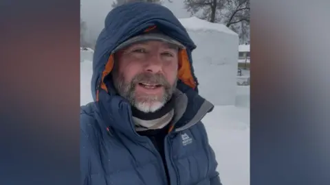 Justin Scott A man is taking a selfie wearing a blue coat with a hood over his head and a cap. He has a grey moustache and short stubble. In the background you can see snow and winter trees.