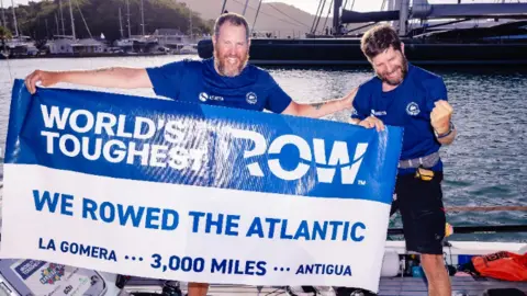 2Stroke.team Karl Austen and Ed Shaw smiling at the camera, wearing navy blue t-shirts and are holding a sign that reads "World's Toughest Row - We rowed the Atlantic".