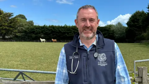 A man with grey hair and a beard is smiling at the camera. He is wearing a checked blue shirt and a navy gilet, which is branded for the vets' practice 'Celtic Equine Veterinary Practice'. He is wearing a stethoscope around his neck. Behind him, there are two horses in the field.