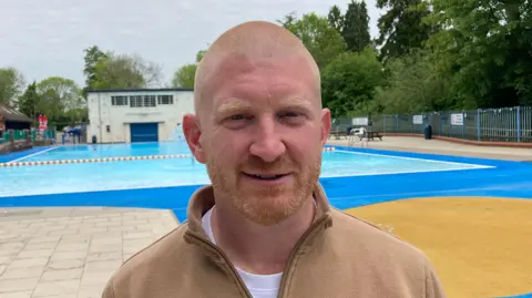 A man with a shaved head and blond beard is pictured wearing a brown fleece while standing in front of an outside swimming pool.