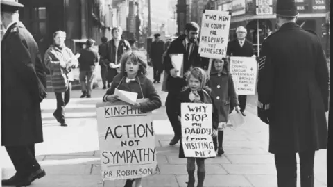 Getty Images A black and white photo of a demonstration. Children are holding signs reading: "King Hill action not sympathy Mr Robinson", "Why stop my Daddy from visiting me?" and "Two men in court for helping the homeless!". Behind them a man's sign reads: "Tory council + Labour government. Broken families at King Hill." A policeman is watching on. There is a double decker bus in the background. 