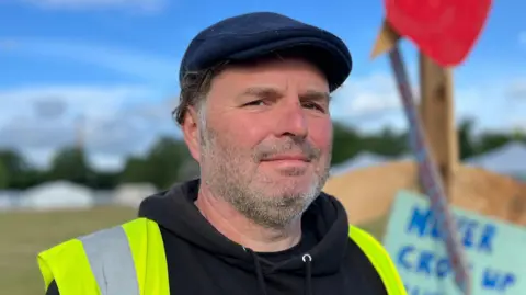 Tom Jackson/BBC Mark Evans is wearing a black jumper, yellow high-vis jacket and a navy cap. He is standing next to a post which has a strawberry and sign attached to it.