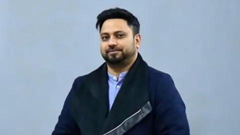 Masum Miah, wearing a dark blue coat, light blue shirt, and jeans, posing with his hands in his pockets against a neutral backdrop.