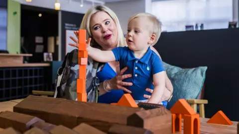 A woman with blonde hair sits at a table and holds a blonde haired toddler on her lap. The boy is stacking up orange building bricks in front of wooden models of buildings on the Elsecar Heritage Centre site.
