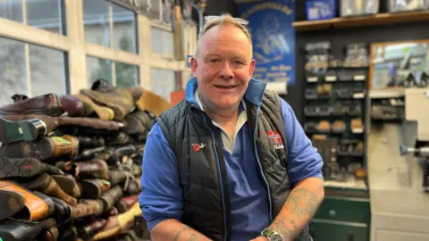 BBC A photograph of a middle-aged man, smiling at the camera. He is white, with short light hair and tattoos on his arms, wearing a black gilet on top of a blue long-sleeved top. He sits in a shop, with shoes piled up in front of a window and tool shelving on the wall.