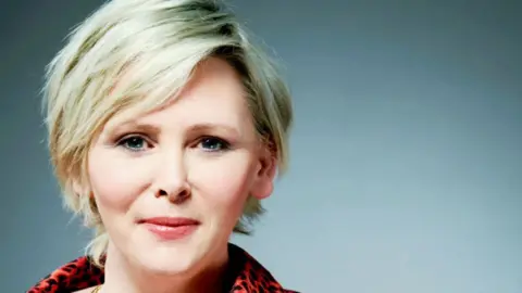 Michelle has short blonde hair and is wearing a red top with black leopard print on it, and two gold necklaces. 