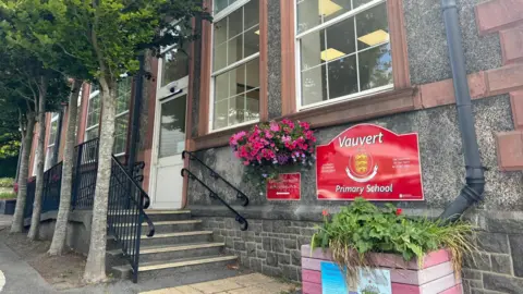 Vauvert Primary School. A victorian school building with a line of trees running next to some steps to the entrance. A red sign with the school's name on.