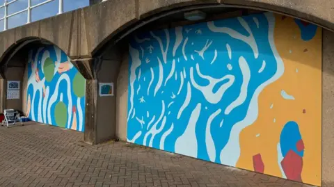 There are two brightly coloured murals in arches under a brown wall.
Both paintings have abstract designs with blue, green and yellow.