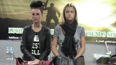Peruvian National Police via Reuters A younger Michaela McCollum, with dark hair in a large bun, stands holding her hands behind her suitcase in Lima Airport. Beside her is Melissa Reid, who has longer blonde hair and is looking at the camera with a neutral expression on her face.