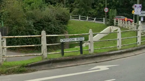 A street sign saying 'Croydon Road'. There is an area of grass behind some railings, and a road. 