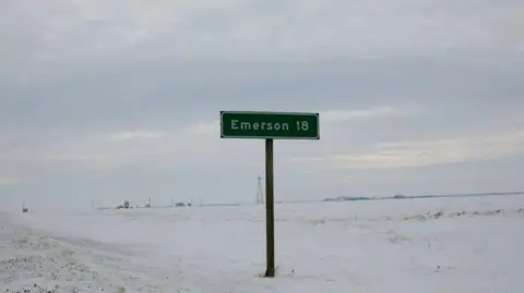 A road sign that says "Emerson 18" in the middle of the snowy field 