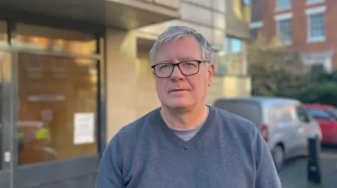 Simon Thake/BBC Tim Renshaw, who has grey hair and black glasses. He is wearing a grey sweater.
