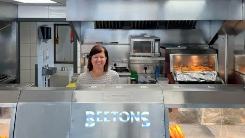 Kim Ashplant stood behind the counter of the fish and chip shop. She is next to the friers and some of the hot food areas shoping the prepared food.
