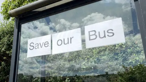 A glass panel at a bus stop with the words "save our bus" printed out on paper and stuck on, there is a hedge behind.