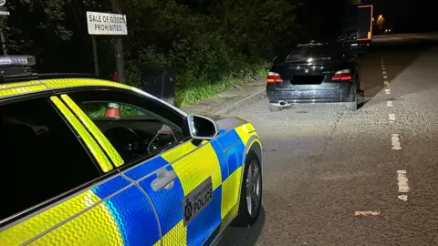 It is night time and there is a Warwickshire Police car on the left, which is neon yellow and blue. In front of it, parked on the road is a black BMW with the license plate blacked out.