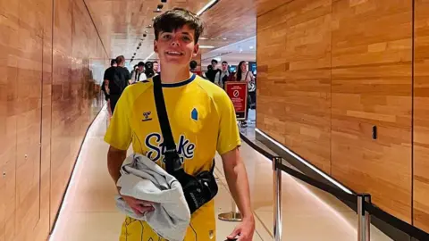 Family photo Mackenzie has short brown hair and is wearing a yellow Everton shirt and a cross body bag. He is stood inside Sydney airport.