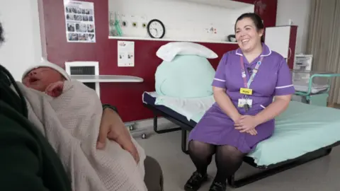 A midwife named Holly, wearing a purple uniform with a yellow name badge, sits on a hospital bed in a maternity ward. She is smiling while speaking, as a newborn baby wrapped in a white blanket is cradled in the arms of a person partially visible in the foreground.