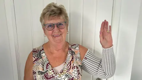 BOB ICB  Gilly Windle is stood in front of a white door and is waving at the camera, smiling. She is wearing a wedding ring and has short, light hair and rimmed glasses.