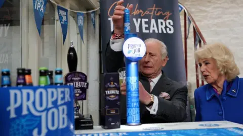 PA King Charles is watched by Queen Camilla as he pulls a pint of beer from a blue pump labelled Proper Job.